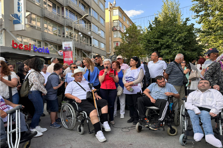 Görselde, açık alanda düzenlenen bir etkinlikte kalabalık bir grup insan bir araya gelmiş. Ön planda tekerlekli sandalyedeki bireyler yer alıyor; bir kadın mikrofonla konuşma yapıyor. Etrafta çeşitli yaş gruplarından insanlar dikkatle dinliyor. Katılımcıların bazıları “Engelliler için eşit haklar” gibi sloganlar içeren tişörtler giymiş. Arka planda bir balık restoranı ve apartmanlar görülüyor.