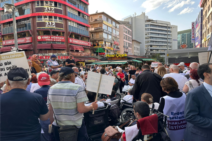 Görselde, kalabalık bir grup insan şehir merkezinde bir araya gelmiş. Aralarında tekerlekli sandalye kullanan bireylerin de bulunduğu grup, ellerinde çeşitli pankartlar taşıyor. Pankartlarda “Engelli Sesleniyor: Haklarımızı İstiyoruz” gibi ifadeler yer alıyor.