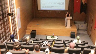 Görselde, bir konferans salonunda düzenlenen bir sunum görülüyor. Kürsüde bir kadın konuşmacı sunum yapıyor; arkasındaki ekranda, engelli bireylerin sosyal hayata katılımı ve istihdamına dair bilgiler yer alıyor. Salonda çeşitli yaş gruplarından kadınlar dinleyici olarak bulunuyor. Sağda Türk bayrağı ve Atatürk portresi, solda ise Aile ve Sosyal Hizmetler Bakanlığı’na ait bir flama yer almakta. Sahne kenarında bir engelli asansörü detayı da dikkat çekiyor.