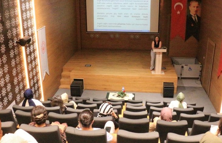 Görselde, bir konferans salonunda düzenlenen bir sunum görülüyor. Kürsüde bir kadın konuşmacı sunum yapıyor; arkasındaki ekranda, engelli bireylerin sosyal hayata katılımı ve istihdamına dair bilgiler yer alıyor. Salonda çeşitli yaş gruplarından kadınlar dinleyici olarak bulunuyor. Sağda Türk bayrağı ve Atatürk portresi, solda ise Aile ve Sosyal Hizmetler Bakanlığı’na ait bir flama yer almakta. Sahne kenarında bir engelli asansörü detayı da dikkat çekiyor.