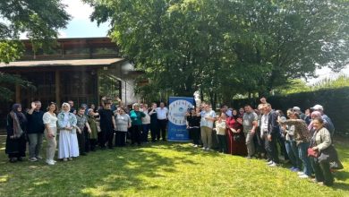 Görselde, yemyeşil ağaçlarla çevrili açık bir alanda geniş bir insan grubu görülüyor. Grup, ortada bulunan mavi bir afişin etrafında toplanmış. Afiş üzerinde "Sakarya Büyükşehir Belediyesi" yazısı ve çeşitli figürler yer alıyor. Katılımcılar farklı yaş gruplarından; bazıları engelli bireyler, bazıları aile üyeleri ya da görevliler gibi görünüyor. Çoğu gülümsüyor ve etkinlikten keyif aldığı belli. Arka planda ahşap bir bina ve bol yapraklı ağaçlar dikkat çekiyor.