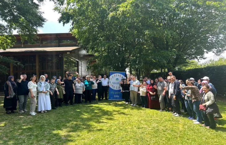 Görselde, yemyeşil ağaçlarla çevrili açık bir alanda geniş bir insan grubu görülüyor. Grup, ortada bulunan mavi bir afişin etrafında toplanmış. Afiş üzerinde "Sakarya Büyükşehir Belediyesi" yazısı ve çeşitli figürler yer alıyor. Katılımcılar farklı yaş gruplarından; bazıları engelli bireyler, bazıları aile üyeleri ya da görevliler gibi görünüyor. Çoğu gülümsüyor ve etkinlikten keyif aldığı belli. Arka planda ahşap bir bina ve bol yapraklı ağaçlar dikkat çekiyor.