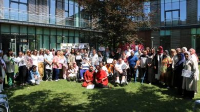 Bu fotoğraf, bir bahçede toplanmış büyük ve neşeli bir kalabalığı gösteriyor. Arkada modern cam cepheli bir bina, önde ise yeşil çimenler üzerinde poz veren yaklaşık 50 kişilik bir grup var. Kalabalığın çoğunluğunu kadınlar oluşturuyor, çeşitli yaşlardan insanlar bir arada. Aralarında engelli bireyler de var; tekerlekli sandalyede ya da bastonla duranlar göze çarpıyor. Bazı kişiler ellerinde sertifika ya da karne tutuyor.