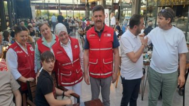 Görselde, bir kafeterya ya da restoran ortamında çekilmiş bir toplu fotoğraf yer alıyor. Ön planda kırmızı Kızılay yelekleri giymiş gönüllüler, bir masa etrafında engelli birey ve yakınlarıyla birlikte poz veriyor. Ortam samimi, neşeli ve kalabalık. Arkada başka masalarda oturan insanlar da görünüyor. Fotoğraf, Türk Kızılayı’nın düzenlediği sosyal bir etkinlikte çekilmiş gibi duruyor; dayanışma, birliktelik ve kapsayıcılık mesajı taşıyor.
