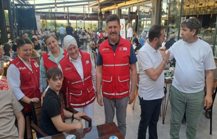 Görselde, bir kafeterya ya da restoran ortamında çekilmiş bir toplu fotoğraf yer alıyor. Ön planda kırmızı Kızılay yelekleri giymiş gönüllüler, bir masa etrafında engelli birey ve yakınlarıyla birlikte poz veriyor. Ortam samimi, neşeli ve kalabalık. Arkada başka masalarda oturan insanlar da görünüyor. Fotoğraf, Türk Kızılayı’nın düzenlediği sosyal bir etkinlikte çekilmiş gibi duruyor; dayanışma, birliktelik ve kapsayıcılık mesajı taşıyor.
