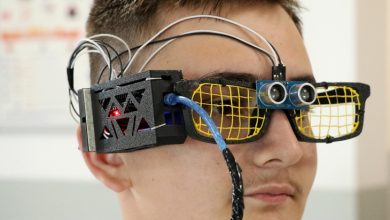 Görselde, bir lise öğrencisi tarafından tasarlanmış teknolojik bir gözlük görülüyor. Gözlüğün ön kısmında sarı ızgaralı camlar ve ultrasonik sensör yer almakta. Üst kısmından geçen kablolar ve yan tarafta bulunan elektronik kutu, cihazın bir prototip olduğunu gösteriyor. Gözlük, görme engelli bireyler için mesafe algılayan bir sistemle tasarlanmış gibi görünüyor.