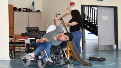 Bu fotoğrafta, spor salonu benzeri kapalı bir mekânda, engelli bir birey tekerlekli sandalyede bocce sporu yaparken görülüyor. Sandalyede yana doğru eğilmiş, kolunu yukarı kaldırmış, yüzünde hem yoğun bir odaklanma hem de kararlılık var. Önünde bocce topunu yuvarlamak için kullanılan özel bir rampa yer alıyor. Arkada bir kadın yardımcı oluyor gibi görünüyor.