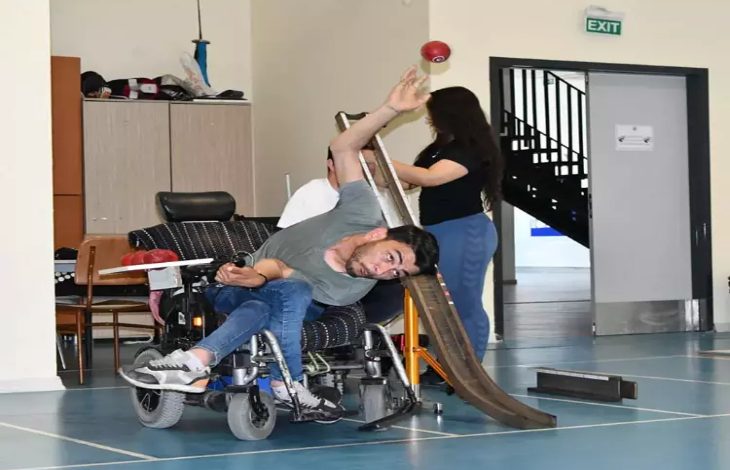 Bu fotoğrafta, spor salonu benzeri kapalı bir mekânda, engelli bir birey tekerlekli sandalyede bocce sporu yaparken görülüyor. Sandalyede yana doğru eğilmiş, kolunu yukarı kaldırmış, yüzünde hem yoğun bir odaklanma hem de kararlılık var. Önünde bocce topunu yuvarlamak için kullanılan özel bir rampa yer alıyor. Arkada bir kadın yardımcı oluyor gibi görünüyor.