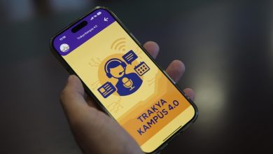 Görselde, bir kişinin elinde tuttuğu akıllı telefonda “TRAKYA KAMPÜS 4.0” adlı bir mobil uygulama açık. Uygulamanın arayüzünde kulaklık takmış bir kişi simgesi, konuşma balonları, mikrofon ve ekran ikonları yer alıyor. Arka plan sarı ve turuncu tonlarında, üst kısmında mor bir başlık çubuğu bulunmakta.