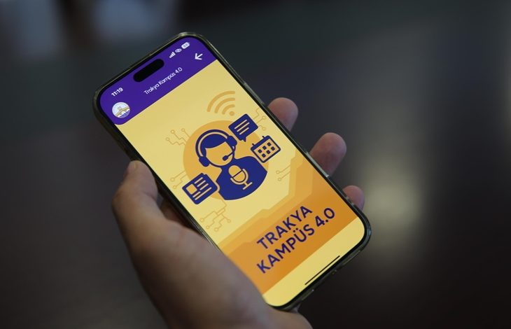 Görselde, bir kişinin elinde tuttuğu akıllı telefonda “TRAKYA KAMPÜS 4.0” adlı bir mobil uygulama açık. Uygulamanın arayüzünde kulaklık takmış bir kişi simgesi, konuşma balonları, mikrofon ve ekran ikonları yer alıyor. Arka plan sarı ve turuncu tonlarında, üst kısmında mor bir başlık çubuğu bulunmakta.