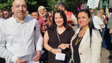 Görselde, açık havada düzenlenen bir etkinlikte üç kişi ön planda poz veriyor. Arka planda ise çeşitli dövizler taşıyan kalabalık bir grup bulunuyor. Öndeki kişilerden biri elinde kâğıt tutuyor, diğer ikisi ise gülümsüyor. Arka plandaki dövizlerden birinde “Sağlık, eğitim ve istihdamda eşit haklarımızı almak istiyoruz” yazısı seçilebiliyor.