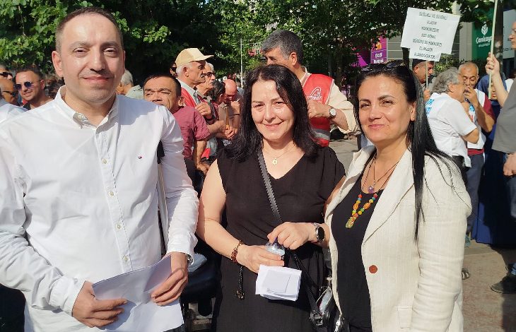 Görselde, açık havada düzenlenen bir etkinlikte üç kişi ön planda poz veriyor. Arka planda ise çeşitli dövizler taşıyan kalabalık bir grup bulunuyor. Öndeki kişilerden biri elinde kâğıt tutuyor, diğer ikisi ise gülümsüyor. Arka plandaki dövizlerden birinde “Sağlık, eğitim ve istihdamda eşit haklarımızı almak istiyoruz” yazısı seçilebiliyor.