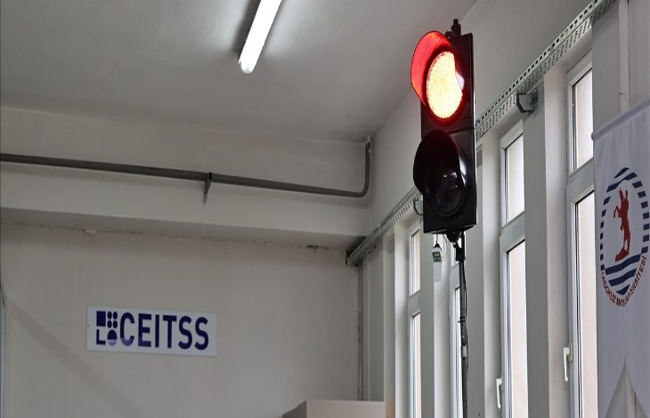 Kapalı bir mekanda trafik lambası yer alıyor. Trafik lambasında kırmızı ışık yanıyor. Lambanın arka planında ise duvarlara asılı bir bayrak ile lambanın tam arkasında bir yazı yer alıyor.