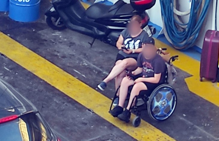 Görselde, bir feribotun araç garajında yer alan iki kişi görülüyor. Biri tekerlekli sandalyede oturan bir çocuk, diğeri ise arkasında oturan ve onu kucaklayan yetişkin bir kadın muhtemelen annesi. İkisi de yazlık giysiler içinde ve telefonlarına bakıyorlar. Etraflarında motosiklet, hortum, bavul ve mavi plastik bidonlar var. Ortam, sıcak ve havasız bir garaj katını andırıyor, zemindeki sarı çizgiler ve kirli beton yüzey dikkat çekiyor.