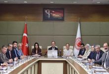 Bu fotoğraf, Türkiye Büyük Millet Meclisi'nde (TBMM) gerçekleşen resmi bir komisyon toplantısını gösteriyor. Masanın başında yer alan kişi, AK Parti İzmir Milletvekili Mehmet Muharrem Kasapoğlu. Arka planda Türk bayrağı, TBMM bayrağı ve Atatürk portresi yer alıyor. Katılımcılar resmi giyimli, çoğunluğu milletvekili ya da bürokratlardan oluşuyor. Masada belgeler, mikrofonlar, isimlikler ve su bardakları bulunuyor. Genel atmosfer ciddi, resmi ve kurumsal.