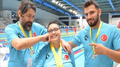 Görselde, bir yüzme havuzu kenarında üç kişi poz veriyor. Üzerlerinde Türk bayrağı armalı mavi tişörtler var ve boyunlarında madalyalar asılı. Ortada duran genç kadın, gözlüklü ve gülümseyerek poz veriyor; hem sağındaki hem solundaki erkekler onunla gurur duyduklarını ifade eden jestler yapıyor. Arkada büyük bir kapalı yüzme havuzu ve renkli bayraklar dikkat çekiyor.