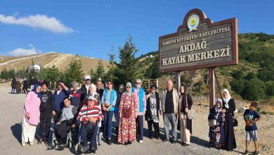 Görselde, Samsun Büyükşehir Belediyesi'ne ait Akdağ Kayak Merkezi tabelasının önünde toplanmış, farklı yaş ve özelliklerde bir grup insan yer almakta. Grup içerisinde tekerlekli sandalye kullanan bireyler, çocuklar, kadınlar ve yaşlılar da bulunuyor. Herkes neşeli, doğayla iç içe bir ortamda olmaktan memnun görünüyor. Arka planda yeşil tepeler, ağaçlar ve açık mavi gökyüzü dikkat çekiyor.