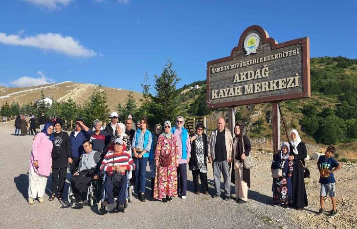Görselde, Samsun Büyükşehir Belediyesi'ne ait Akdağ Kayak Merkezi tabelasının önünde toplanmış, farklı yaş ve özelliklerde bir grup insan yer almakta. Grup içerisinde tekerlekli sandalye kullanan bireyler, çocuklar, kadınlar ve yaşlılar da bulunuyor. Herkes neşeli, doğayla iç içe bir ortamda olmaktan memnun görünüyor. Arka planda yeşil tepeler, ağaçlar ve açık mavi gökyüzü dikkat çekiyor.