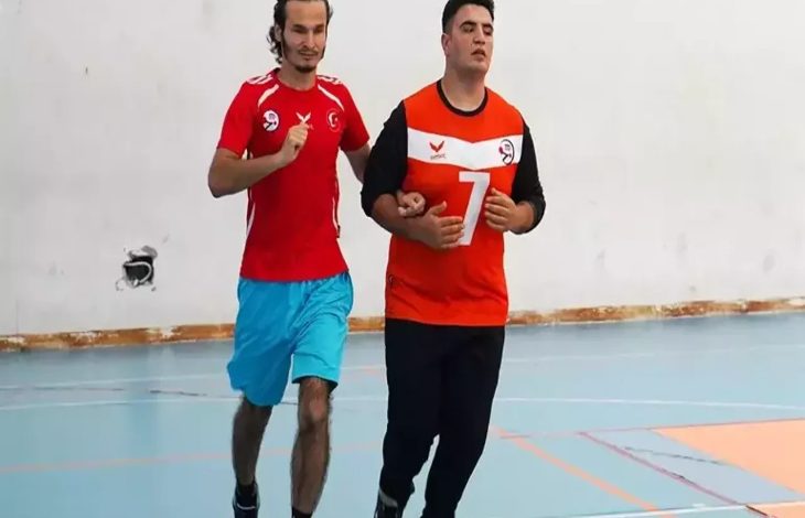Görselde bir spor salonunda iki genç erkek yan yana koşuyor. Sol taraftaki kişi mavi şort ve kırmızı tişört giymiş; sağ taraftaki ise turuncu-siyah forma giyiyor ve üzerinde "7" numarası yer alıyor. Sağdaki bireyin gözleri kapalı görme engelli olduğu anlaşılıyor; diğer kişi ona rehberlik ediyor. İkili, antrenman ya da sportif bir etkinlik sırasında uyumlu şekilde hareket ediyor. Arka plan sade bir duvar ve spor salonu zeminiyle çevrili.