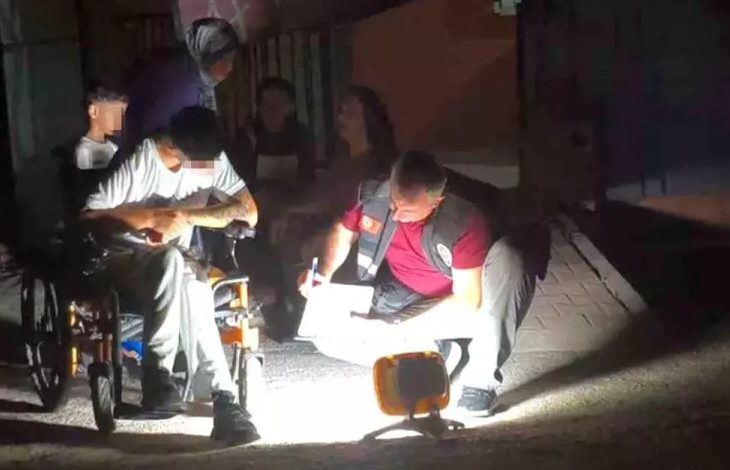 Bu görselde, gece vakti dış mekânda gerçekleşen bir olayın ardından bir polis memuru, tekerlekli sandalyedeki bir bireyden ifade alıyor. Aydınlatma taşınabilir bir ışıkla sağlanmış ve sahneye yoğun bir ışık tutulmuş. Polis memuru yerde diz çökerek not alırken, engelli birey olayla ilgili bilgi veriyor. Arka planda başka kişiler de bulunuyor.