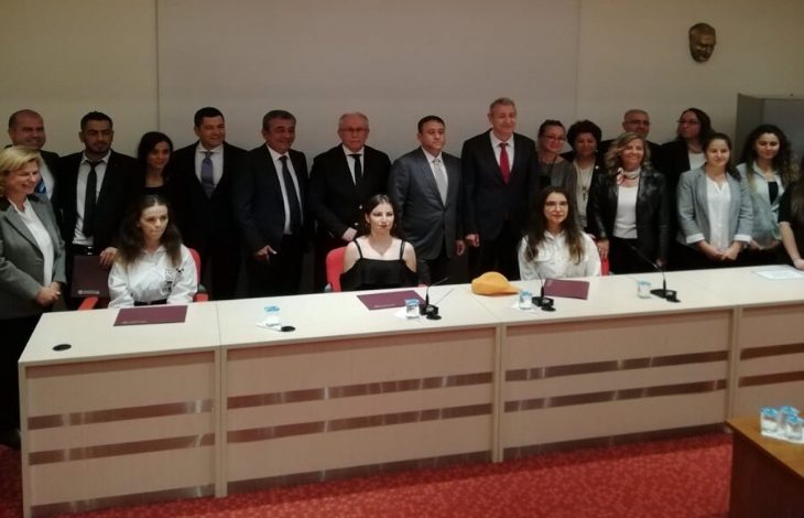 Görselde, bir masanın arkasında oturan üç kadın yer alıyor. Kadınların arkasında ise, ayakta duran kadın ve erkeklerden oluşan kalabalık bir grup bulunuyor.