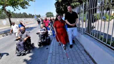 Görselde, bir grup engelli birey ve onlara eşlik eden kişiler bir kaldırım üzerinde yürürken görülüyor. Önde kırmızı elbiseli bir kadın beyaz bastonuyla ilerliyor; yanında ona eşlik eden bir adam var. Arkalarında akülü tekerlekli sandalye kullanan bireyler ve diğer katılımcılar bulunuyor. Ortam gündüz, açık hava ve güneşli bir gün. Görsel, Bornova Belediyesi’nin engellilerin günlük yaşamda karşılaştığı erişilebilirlik sorunlarına dikkat çekmek için düzenlediği bir farkındalık yürüyüşünü yansıtıyor.