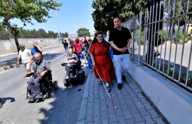 Görselde, bir grup engelli birey ve onlara eşlik eden kişiler bir kaldırım üzerinde yürürken görülüyor. Önde kırmızı elbiseli bir kadın beyaz bastonuyla ilerliyor; yanında ona eşlik eden bir adam var. Arkalarında akülü tekerlekli sandalye kullanan bireyler ve diğer katılımcılar bulunuyor. Ortam gündüz, açık hava ve güneşli bir gün. Görsel, Bornova Belediyesi’nin engellilerin günlük yaşamda karşılaştığı erişilebilirlik sorunlarına dikkat çekmek için düzenlediği bir farkındalık yürüyüşünü yansıtıyor.