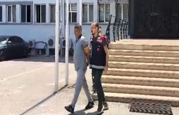 Görselde, elleri arkadan kelepçelenmiş bir suçlu ile bir polis memuru yan yana yürüyor. Polis memuru, suçlunun kaçmaması için onun kolunu tutuyor.