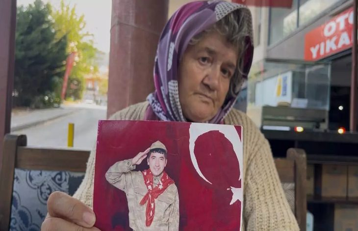 Fotoğrafta yaşlı bir kadın, duygusal bir ifadeyle elinde genç bir askerin fotoğrafını tutuyor. Kadın başörtülü, hüzünlü ve yorgun bir yüz ifadesine sahip. Tuttuğu fotoğrafta, Türk bayrağı fonu önünde selam veren bir genç asker yer alıyor. Mekân, bir sokak kenarındaki açık bir oturma alanı gibi görünüyor; arka planda bir "oto yıkama" tabelası dikkat çekiyor.