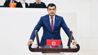 Görselde, TBMM Genel Kurulu’nda bir erkek milletvekili kürsüde konuşma yaparken görülüyor. Üzerinde koyu mavi bir takım elbise ve kırmızı desenli bir kravat bulunuyor. Kürsüde TBMM logosu yer alıyor ve dijital ekranda “00:19” süresi görünüyor. Arkada oturan dört kişi daha var; biri beyaz başörtülü bir kadın, diğerleri erkek ve takım elbise giymiş.