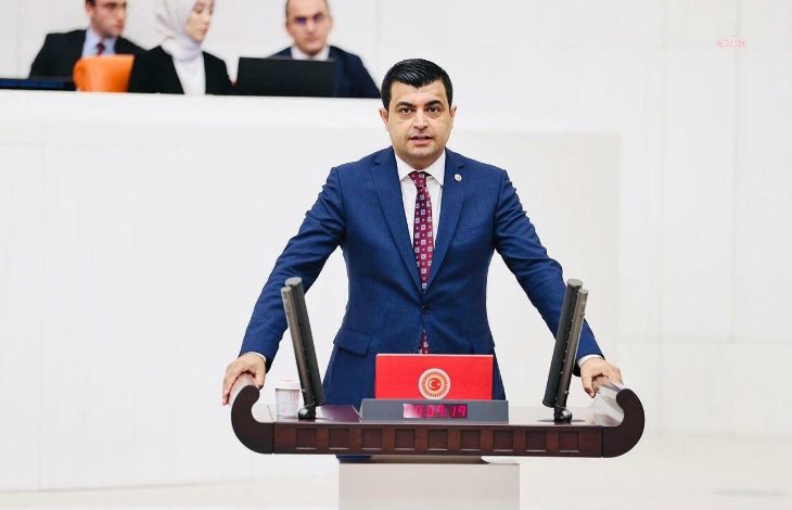 Görselde, TBMM Genel Kurulu’nda bir erkek milletvekili kürsüde konuşma yaparken görülüyor. Üzerinde koyu mavi bir takım elbise ve kırmızı desenli bir kravat bulunuyor. Kürsüde TBMM logosu yer alıyor ve dijital ekranda “00:19” süresi görünüyor. Arkada oturan dört kişi daha var; biri beyaz başörtülü bir kadın, diğerleri erkek ve takım elbise giymiş.