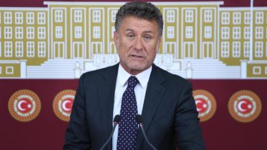 Görselde, takım elbise giymiş bir erkek siyasetçi, Türkiye Büyük Millet Meclisi (TBMM) amblemleri ve meclis binası illüstrasyonu önünde basın açıklaması yapıyor. Önünde iki mikrofon yer alıyor. Arka planda kırmızı zemin üzerinde Türk bayrakları ve TBMM logoları görülüyor.