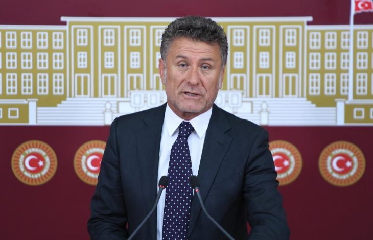 Görselde, takım elbise giymiş bir erkek siyasetçi, Türkiye Büyük Millet Meclisi (TBMM) amblemleri ve meclis binası illüstrasyonu önünde basın açıklaması yapıyor. Önünde iki mikrofon yer alıyor. Arka planda kırmızı zemin üzerinde Türk bayrakları ve TBMM logoları görülüyor.