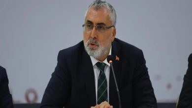 Görselde, takım elbise giymiş, kravat takan ve gözlüklü bir erkek, mikrofon önünde konuşma yaparken görülüyor. Arka planda sade bir fon yer almakta. Masada mikrofon, kâğıtlar ve su bardakları bulunuyor.