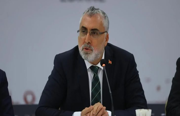 Görselde, takım elbise giymiş, kravat takan ve gözlüklü bir erkek, mikrofon önünde konuşma yaparken görülüyor. Arka planda sade bir fon yer almakta. Masada mikrofon, kâğıtlar ve su bardakları bulunuyor.