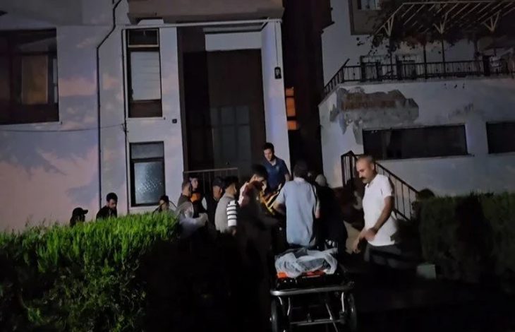 Görselde, gece vakti bir apartman önünde toplanmış bir grup insan görülüyor. Ortam karanlık ve acil bir durum izlenimi veriyor. Ön planda boş bir sedye yer alırken, arka planda sağlık ve kurtarma ekipleri ile bazı vatandaşlar bir kişiye müdahale ediyor gibi görünüyor. Bina eski ve hasarlı bir yapı izlenimi veriyor.