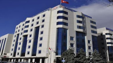 Görselde, Ankara'da bulunan Başkent Üniversitesi Hastanesi’nin modern ve çok katlı ana binası yer alıyor. Bina, beyaz cepheli ve mavi camlı mimarisiyle dikkat çekiyor. Üst kısmında Türk bayrağı ve hastane logosu dalgalanıyor. Ön cephede araçlar park halinde ve giriş kısmı net şekilde görülüyor.
