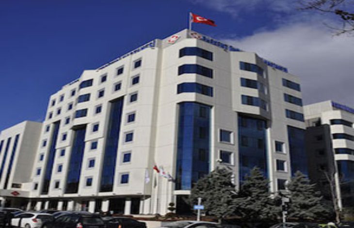 Görselde, Ankara'da bulunan Başkent Üniversitesi Hastanesi’nin modern ve çok katlı ana binası yer alıyor. Bina, beyaz cepheli ve mavi camlı mimarisiyle dikkat çekiyor. Üst kısmında Türk bayrağı ve hastane logosu dalgalanıyor. Ön cephede araçlar park halinde ve giriş kısmı net şekilde görülüyor.