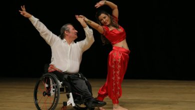 Görselde, sahnede dans eden iki kişi yer alıyor. Sol tarafta tekerlekli sandalye kullanan bir erkek, sağ kolunu zarifçe yukarı kaldırmış şekilde dans ediyor. Yanında ise parlak kırmızı geleneksel tarzda kıyafet giymiş bir kadın, zarif bir dans pozuyla ona eşlik ediyor.