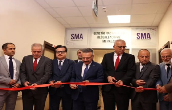 Bu görselde, bir açılış töreni sırasında kırmızı kurdele kesen bir grup erkek görülüyor. Hepsi resmi kıyafetler giymiş; takım elbise ve kravatlı. Arka planda “SMA” ve “Genetik Hastalıklar Değerlendirme Merkezi” yazılı tabela yer almakta.