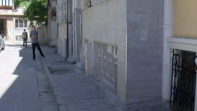 Görselde, dar bir sokakta çekilmiş bir apartman önü yer almakta. Kaldırım kenarında yürüyen iki adam görünüyor, biri önde, diğeri arkada. Sağdaki bina girişinin önünde ferforje bir kapı ve bodrum pencerelerini kapatan demir parmaklıklar mevcut. Işık ve gölge kontrastı, öğle saatlerine yakın bir zamanda çekildiğini düşündürüyor.