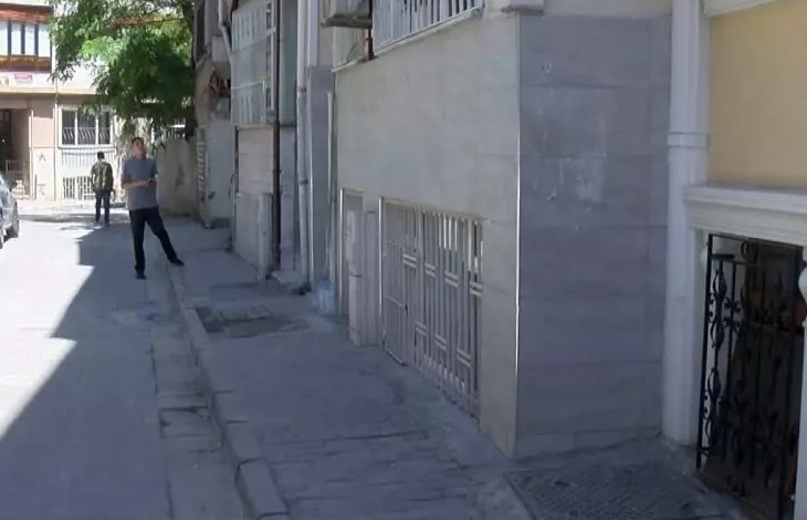 Görselde, dar bir sokakta çekilmiş bir apartman önü yer almakta. Kaldırım kenarında yürüyen iki adam görünüyor, biri önde, diğeri arkada. Sağdaki bina girişinin önünde ferforje bir kapı ve bodrum pencerelerini kapatan demir parmaklıklar mevcut. Işık ve gölge kontrastı, öğle saatlerine yakın bir zamanda çekildiğini düşündürüyor.