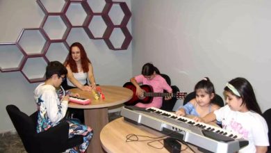 Görselde, bir müzik sınıfında öğretmen ve çocuklardan oluşan bir grup görülüyor. Ortada, kırmızı saçlı bir kadın öğretmen (görme engelli müzik öğretmeni Simay Boz), yuvarlak bir masada oturmuş; etrafında ise farklı enstrümanlarla ilgilenen çocuklar bulunuyor.