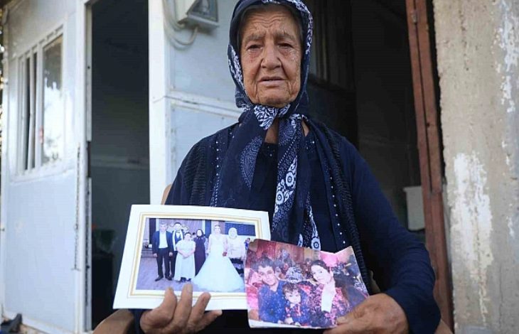 Bu görselde, başörtülü yaşlı bir kadın, elinde iki aile fotoğrafı tutarak kameraya bakıyor. Kadının yüz ifadesi hüzünlü ve derin bir acıyı yansıtıyor. Arka planda mütevazı bir evin girişi görülüyor. Fotoğraflardan biri düğün anına, diğeri ise ailece çekilmiş bir kareye ait.