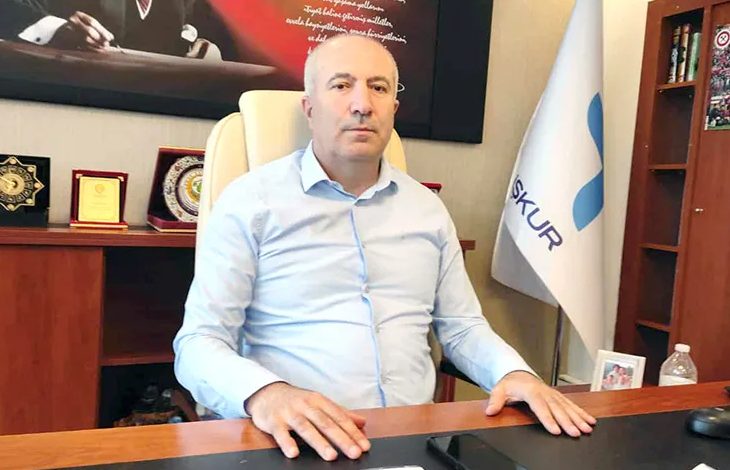 Görselde, bir makam odasında oturan bir erkek yer alıyor. Kendisinin ciddi ve kararlı bir ifadesi var. Üzerinde açık mavi bir gömlek bulunuyor. Masasının arkasında Türkiye İş Kurumu'na (İŞKUR) ait bir bayrak, çeşitli plaketler ve ödüller yer almakta. Sol arka planda Atatürk'ün yer aldığı bir tablo dikkat çekiyor. Masa üzerinde bir su şişesi ve çerçeveli bir fotoğraf da görülüyor.