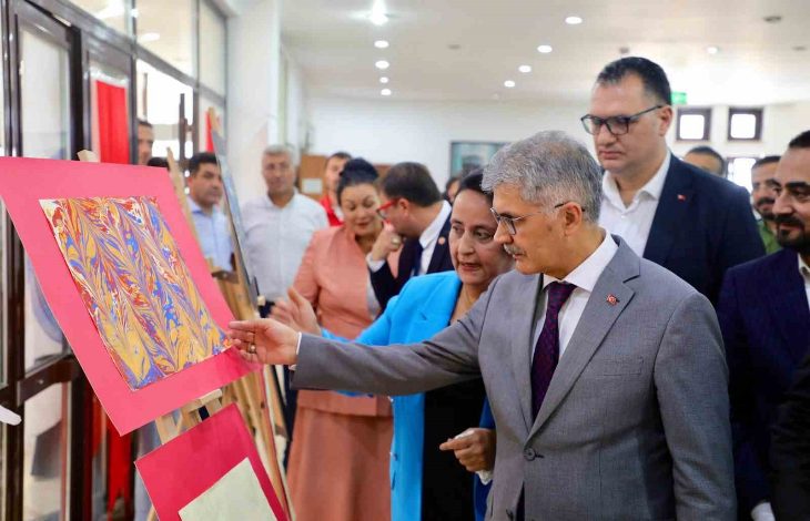 Görselde bir sanat sergisi açılışından bir an yer almakta. Önde takım elbiseli bir adam (muhtemelen bir yetkili ya da protokol üyesi), pembe zemin üzerine yerleştirilmiş geleneksel bir ebru sanat eserine dikkatle bakıyor ve eliyle eseri işaret ediyor. Yanında mavi ceketli bir kadın ve arka planda farklı renklerde giyinmiş birçok kişi daha yer almakta. Katılımcıların resmi giyimli olması, etkinliğin ciddi ve resmi bir açılış olduğunu gösteriyor.