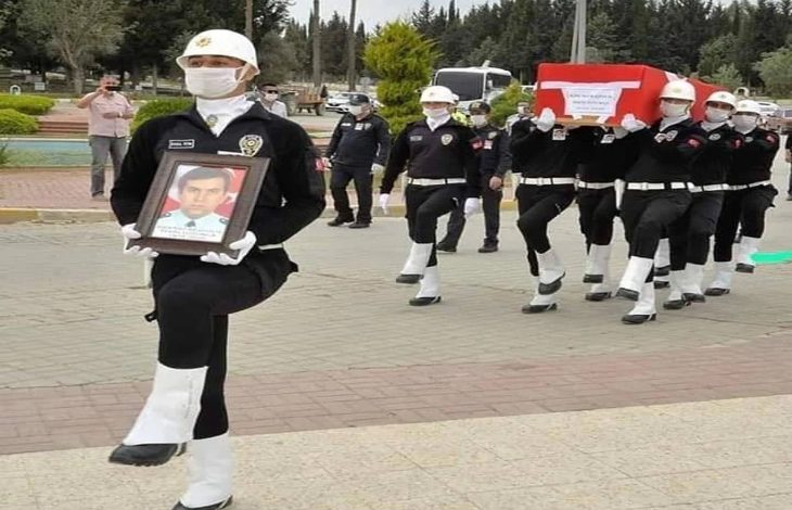 Görselde, cenaze töreni sırasında görev şehidi bir polis memuru için resmi tören düzenleniyor. Önde yürüyen bir polis memuru, hayatını kaybeden meslektaşının fotoğrafını taşıyor. Arkasında ise altı polis memuru, Türk bayrağına sarılı tabutu omuzlarında taşıyor. Hepsi resmi kıyafetli, beyaz kask ve eldiven takmış durumda. Tören ciddiyet ve saygı içinde ilerliyor; çevrede bazı insanlar töreni izliyor. Bu kare, hem hüzün hem de onur dolu bir vedayı yansıtıyor.