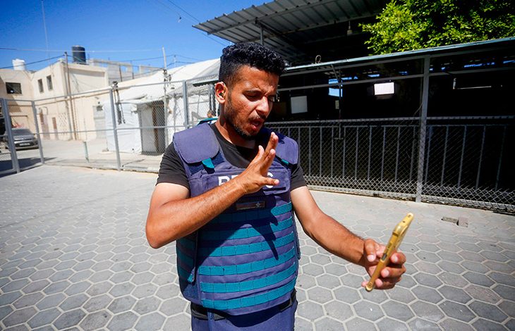 Görselde, Gazze’de bulunan bir erkek aktivist, üzerinde mavi koruyucu basın yeleğiyle cep telefonuna bakıyor ve işaret diliyle iletişim kuruyor. Arka planda basit yapılar, tellerle çevrili bir alan ve açık hava ortamı görülüyor. Güneşli bir günde çekilmiş olan bu karede, kişi kararlı bir ifadeyle dünyaya mesajını iletmeye çalışıyor.