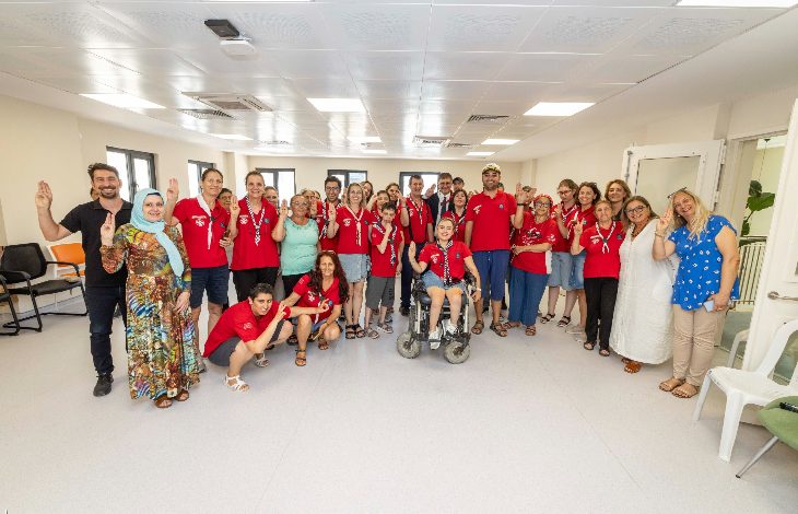 Bu görselde, geniş ve aydınlık bir kapalı mekânda toplanmış kalabalık bir grup insan görülüyor. Çoğu kişi kırmızı tişört giymiş ve boynunda beyaz fular takmış, bu da muhtemelen bir organizasyona veya etkinliğe ait olduklarını gösteriyor. Ortada tekerlekli sandalyede bir birey bulunuyor. Sol köşede geleneksel kıyafetli bir kadın ve sağ köşede mavi puantiyeli bluz giymiş bir kadın yer alıyor.