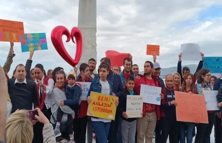 Görselde, ellerinde pankartlar taşıyan bir grup insan açık alanda toplanmış. Pankartlarda otizmli bireylerin anlaşılması, istihdam hakları ve farkındalık taleplerine dair ifadeler yer alıyor. Kalabalık, toplumsal farkındalık ve hak arayışı amacıyla bir araya gelmiş. Arka planda büyük bir kalp figürü ve Türk bayrağı da görülüyor.